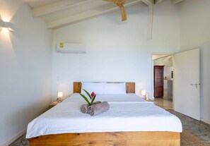 A hotel room with a wooden bed frame, white bedding, a small bedside table with a lamp, and a wall-mounted air conditioner. - Romantic garden bungalow with pool and sea view (Sint Willibrordus)