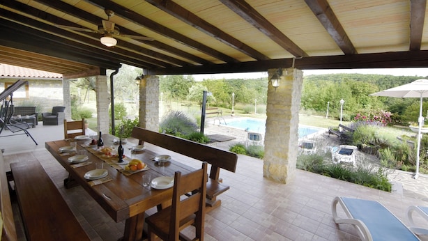 An outdoor dining area with a wooden table set for four, surrounded by stone pillars and a view of a swimming pool and garden. - Villa With Private Pool and Garden Retreat (Groznjan)