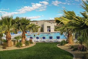 Zona de piscina con sillones de descanso, palmeras y un edificio con un cartel.