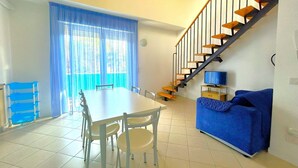 A dining area with a table and chairs, a blue sofa, and a staircase. - COND. PELICAN - A2 7+0 - B80A6A (Bibione Pineda)