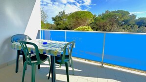 A balcony with a table and chairs, a blue railing, and a view of trees and hills. - COND. PELICAN - A2 7+0 - B80A6A (Bibione Pineda)