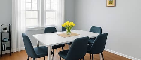 A dining room with a white table, four dark chairs, a vase of yellow flowers, a picture on the wall, and a black shelf with a plant.