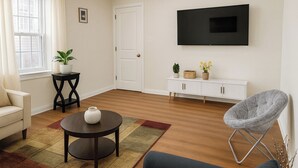 A living room with a sofa, a round table with a vase, a grey chair, a TV on the wall, a white cabinet, and a potted plant. - LGA AIRPORT (Corona)