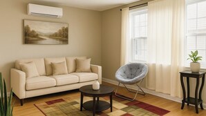 A living room with a beige sofa, a round coffee table, a gray chair, a small table with a plant, and a painting on the wall. - LGA AIRPORT (Corona)