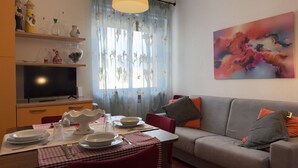 Ein Wohnzimmer mit einer Couch, einem Esstisch gedeckt mit Tellern und Besteck, einem Fernseher und einem Gemälde an der Wand.