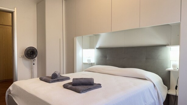 Ein ordentlich bezogenes Bett mit weißer Decke und zwei gefalteten Handtüchern, eine Nachttischlampe, ein Wandventilator und ein großer Spiegel über dem Bett.
