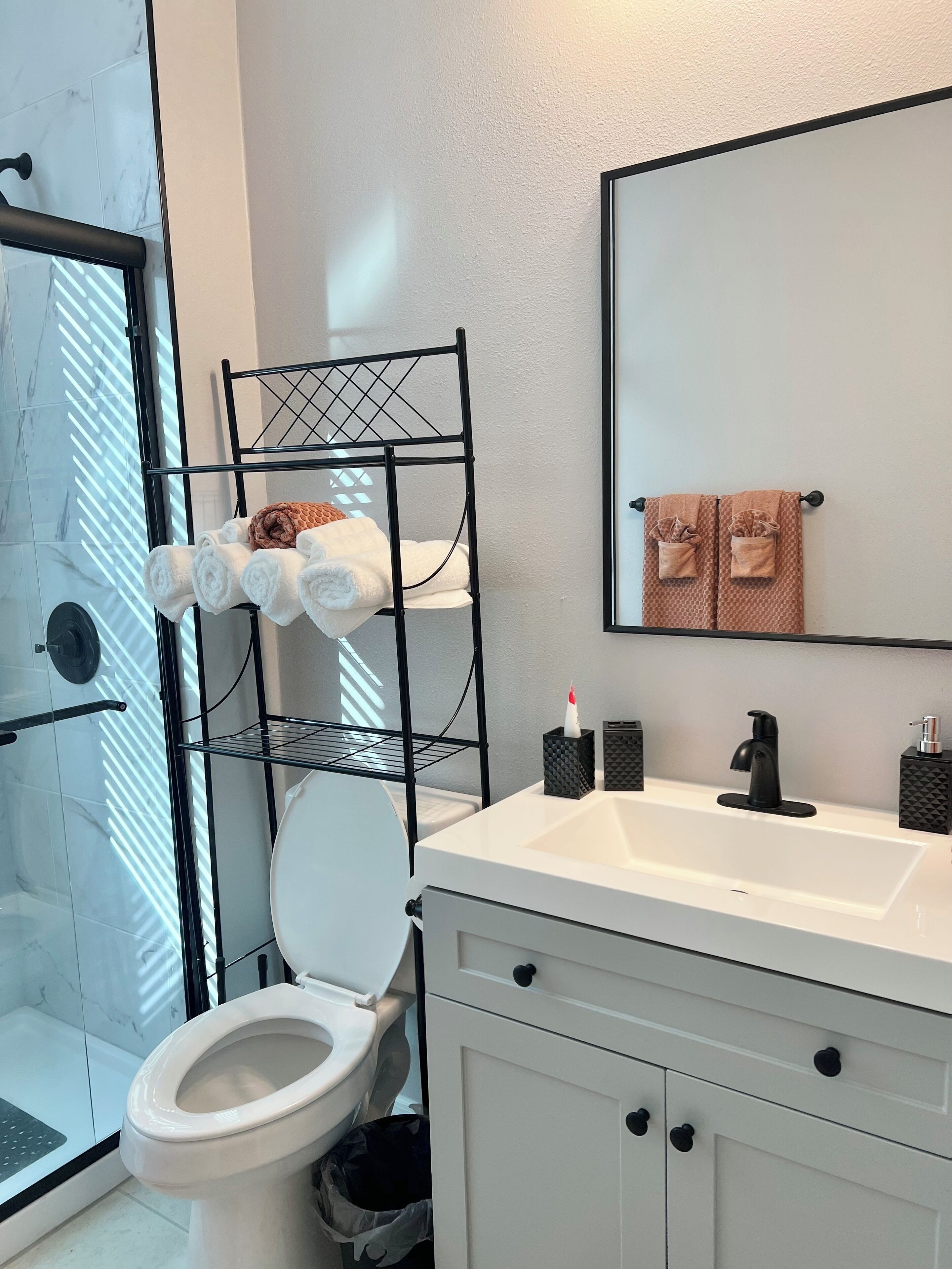 A bathroom with a white sink, black faucet, and a mirror above it. There is a towel rack with towels and a toilet with a white lid.