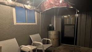 Un espace patio avec deux chaises blanches, un tonneau en métal et une fenêtre dont les stores sont fermés.