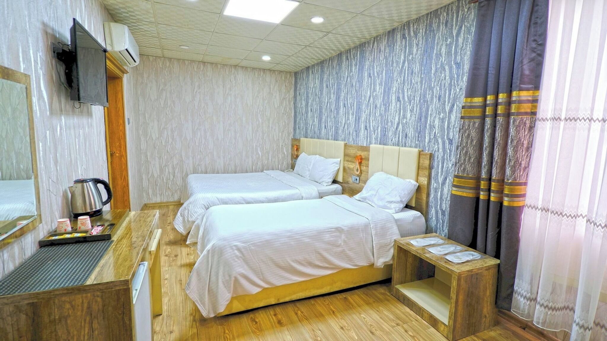 Habitación de hotel con dos camas, un televisor, aire acondicionado, un escritorio de madera con hervidor y una banca de madera.