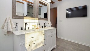 Un baño con un espejo grande, dos lavamanos, un mueble de tocador con cajones, una toalla y un televisor montado.