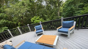 Una terraza con asientos acolchados azules y una mesa de madera, rodeada por barandillas de madera y abundante vegetación.