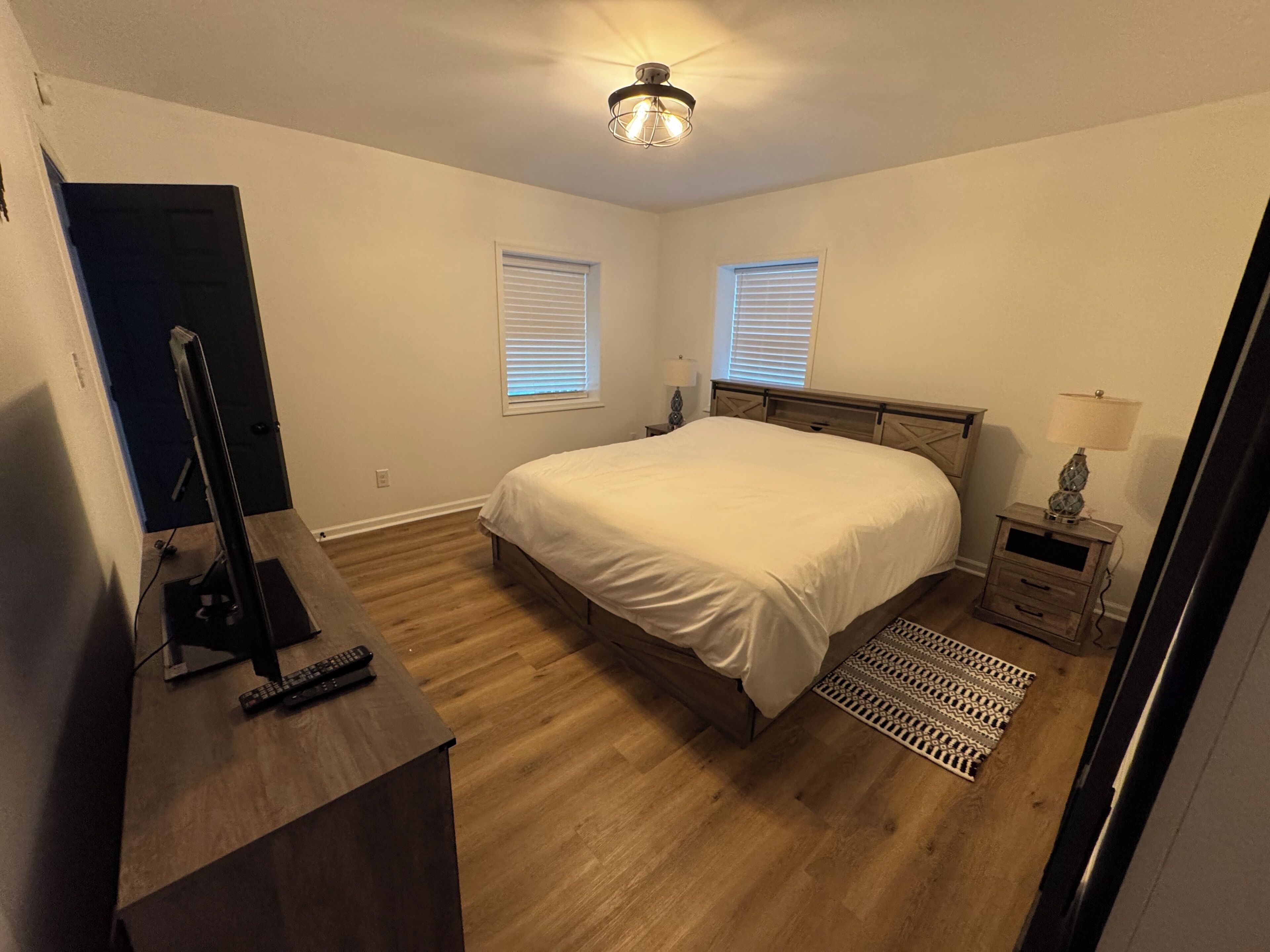 A bedroom with a bed, a desk with a computer and keyboard, two bedside tables with lamps, and a ceiling light.
