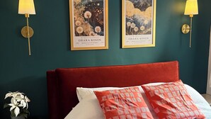 Ein Schlafzimmer mit rotem Kopfteil, weißer und roter Bettwäsche, zwei gerahmten Kunstwerken, einem kleinen Nachttisch mit Lampe und einem Blumentopf.