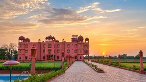 A large pink building with multiple domes and a central entrance, surrounded by a landscaped garden and a paved pathway leading to a pool. - Harigarh Resort (Sumerpur)
