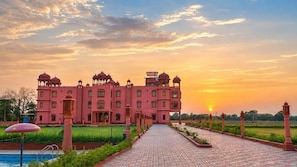 A large pink building with multiple domes and a central entrance, surrounded by a landscaped garden and a paved pathway leading to a pool. - Harigarh Resort (Sumerpur)