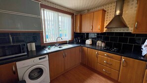A compact kitchen with wooden cabinets, a built-in washing machine, and a window with blinds.