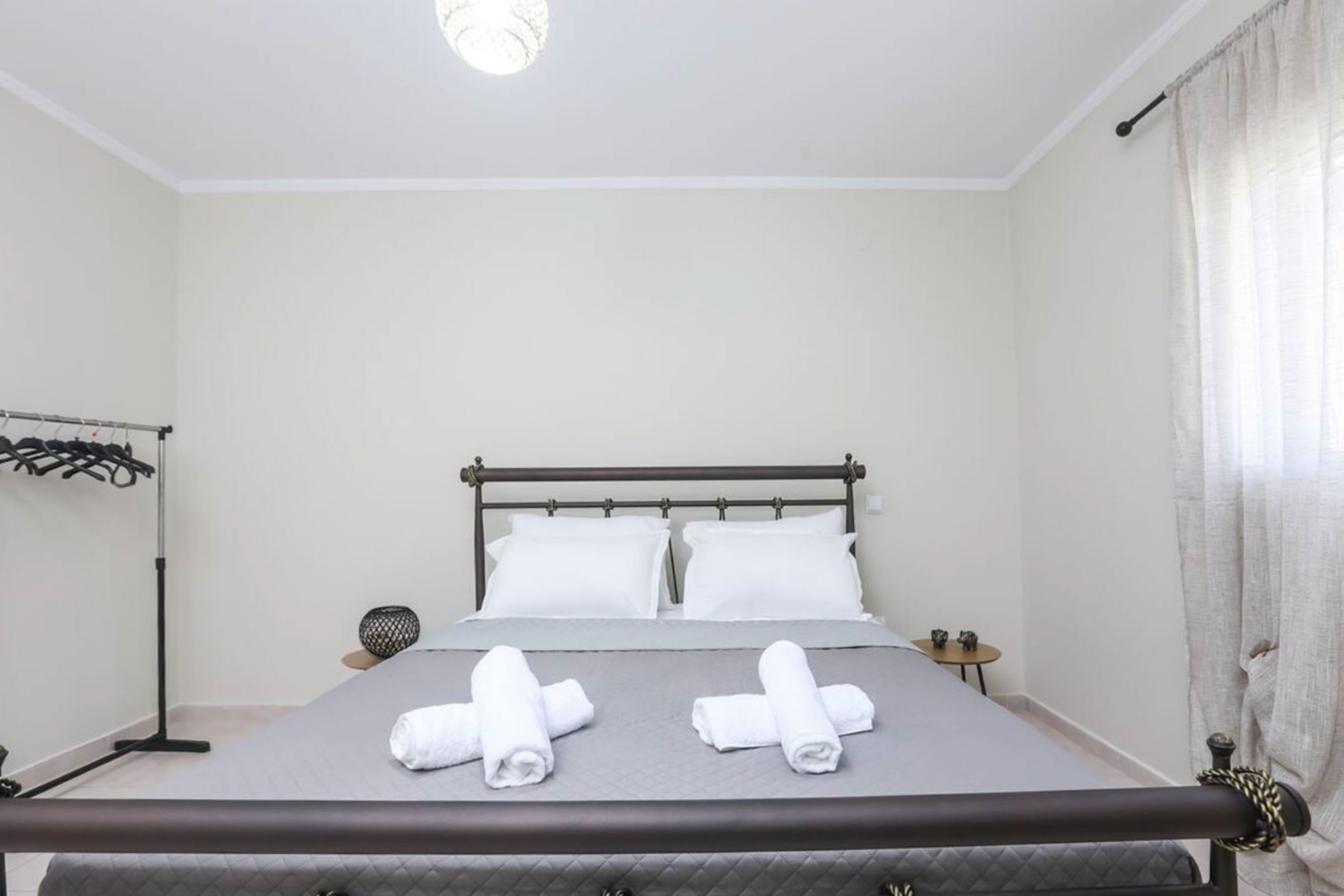Uma cama bem arrumada com duas toalhas enroladas, uma mesa de cabeceira com abajur e um cabideiro na parede.