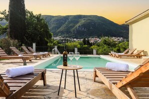 Un espace aménagé au bord de la piscine, avec des chaises longues, une table sur laquelle se trouvent une bouteille et des verres, et une vue sur une ville et des montagnes en arrière-plan.
