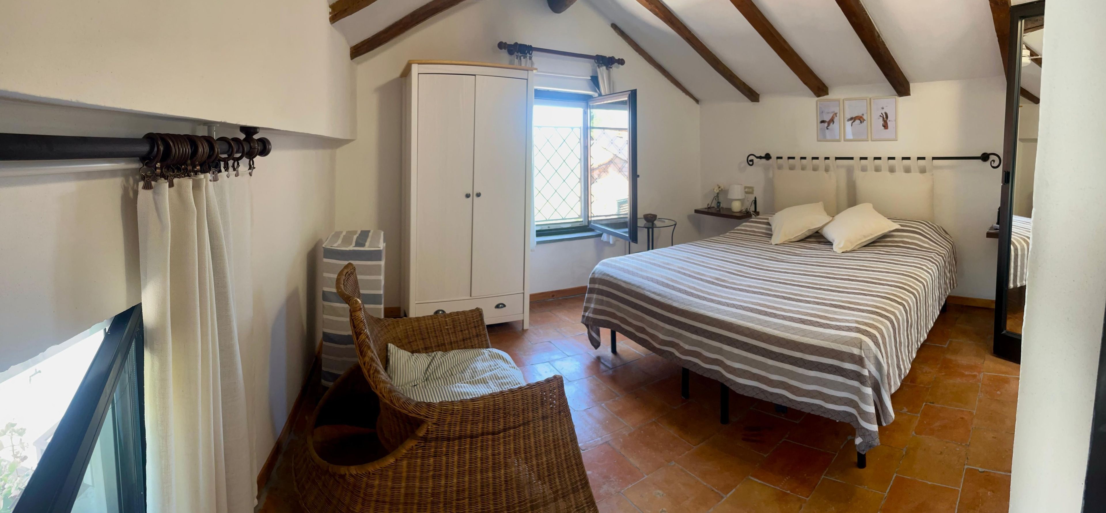 Une chambre avec un lit, une armoire, un fauteuil en osier et une fenêtre donnant sur un beau paysage.