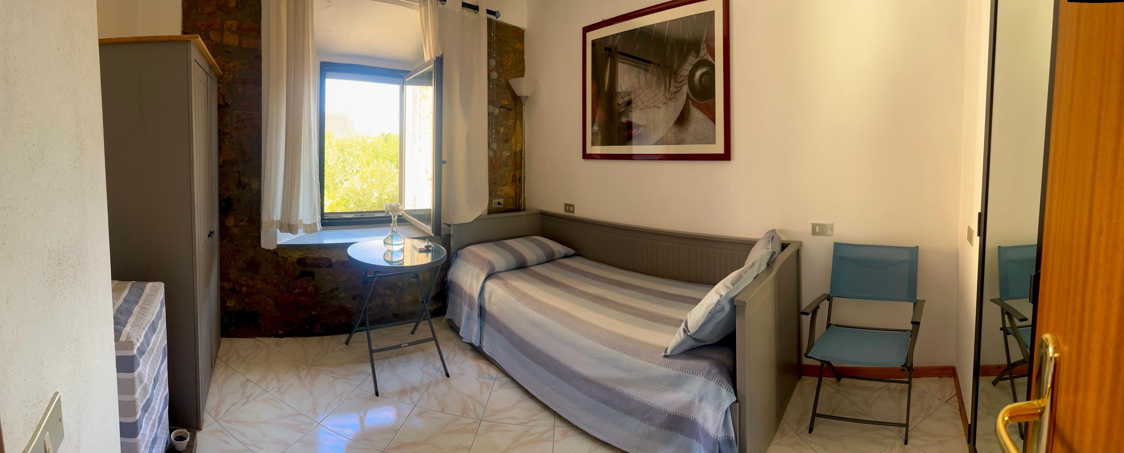 Una stanza piccola e funzionale con un letto, un tavolo e una sedia. Presenta una finestra con tende, un quadro appeso al muro e una porta.