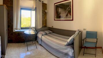 Una stanza piccola e funzionale con un letto, un tavolo e una sedia. Presenta una finestra con tende, un quadro appeso al muro e una porta.