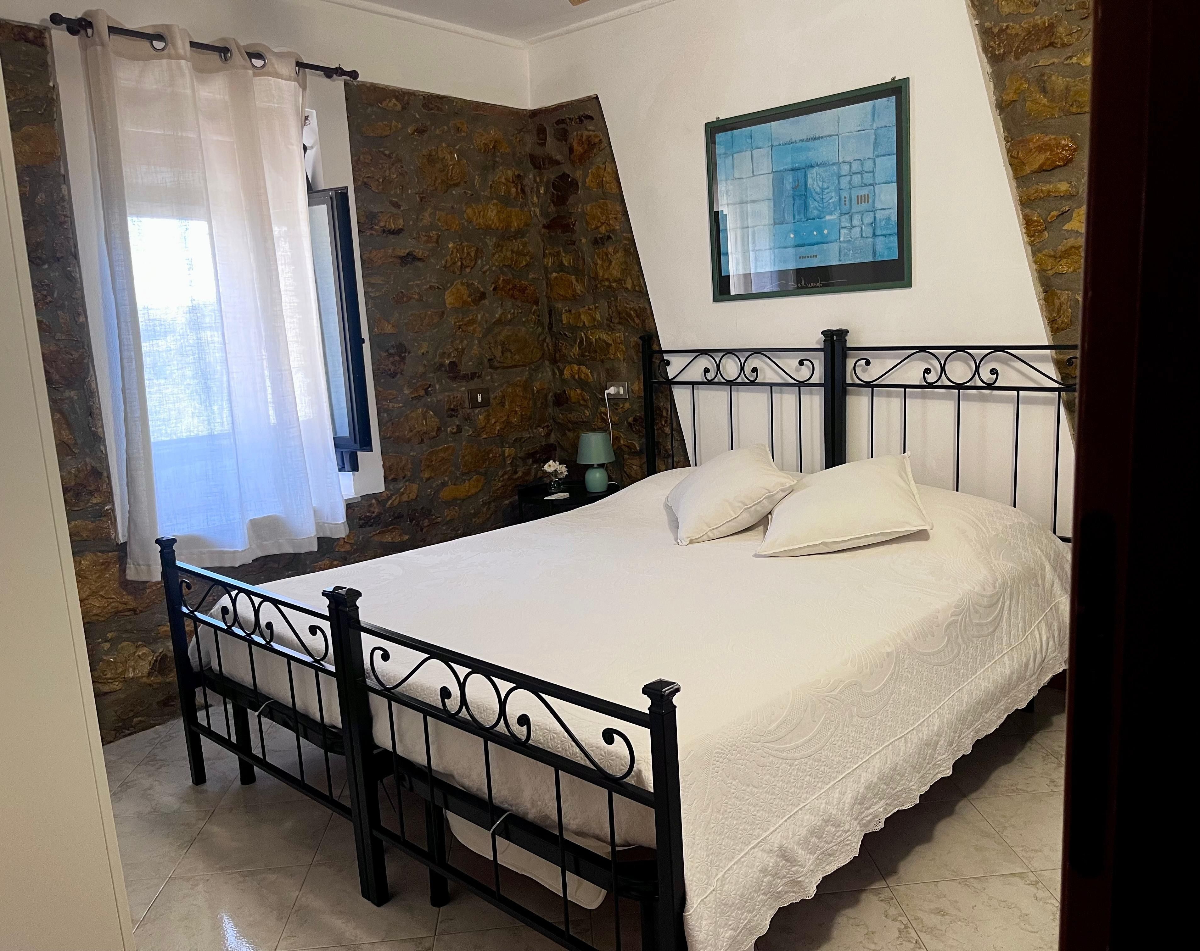 A bedroom with a stone wall, a bed with a metal frame, a window with a curtain, and a painting on the wall.