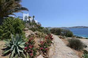 A coastal building with a stone pathway, surrounded by greenery and overlooking the sea. - Topos exclusive Mykonos- Villa Poseidon (Agios Ioannis Diakoftis)