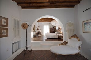 Une pièce avec un lit blanc, un fauteuil blanc, un plafond en bois, une cheminée et une grande porte en arc de cercle.