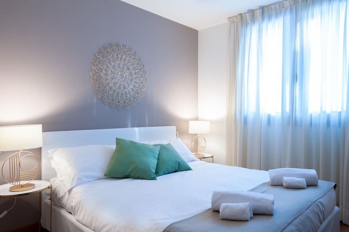 Een net opgemaakt bed met kussens, een kunstwerk aan de muur en een raam met gordijnen.