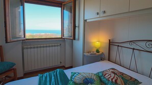 Ein Schlafzimmer mit Bett, Fenster mit Meerblick, Heizkörper, Nachttisch mit Lampe und einem Sessel.
