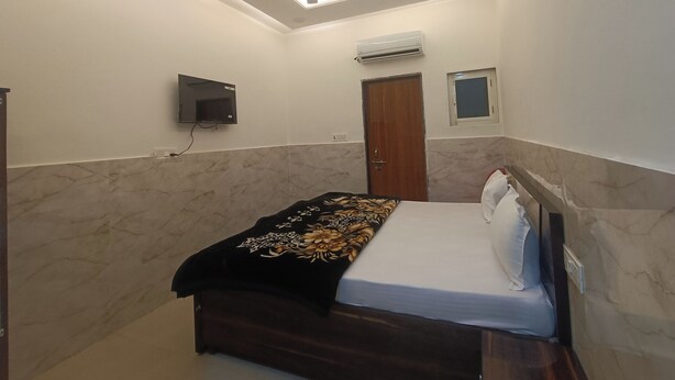 A bedroom with a bed, a ceiling fan, an air conditioning unit, a TV, and a door. - Hotel Krishnam Palace (Vrindavan)