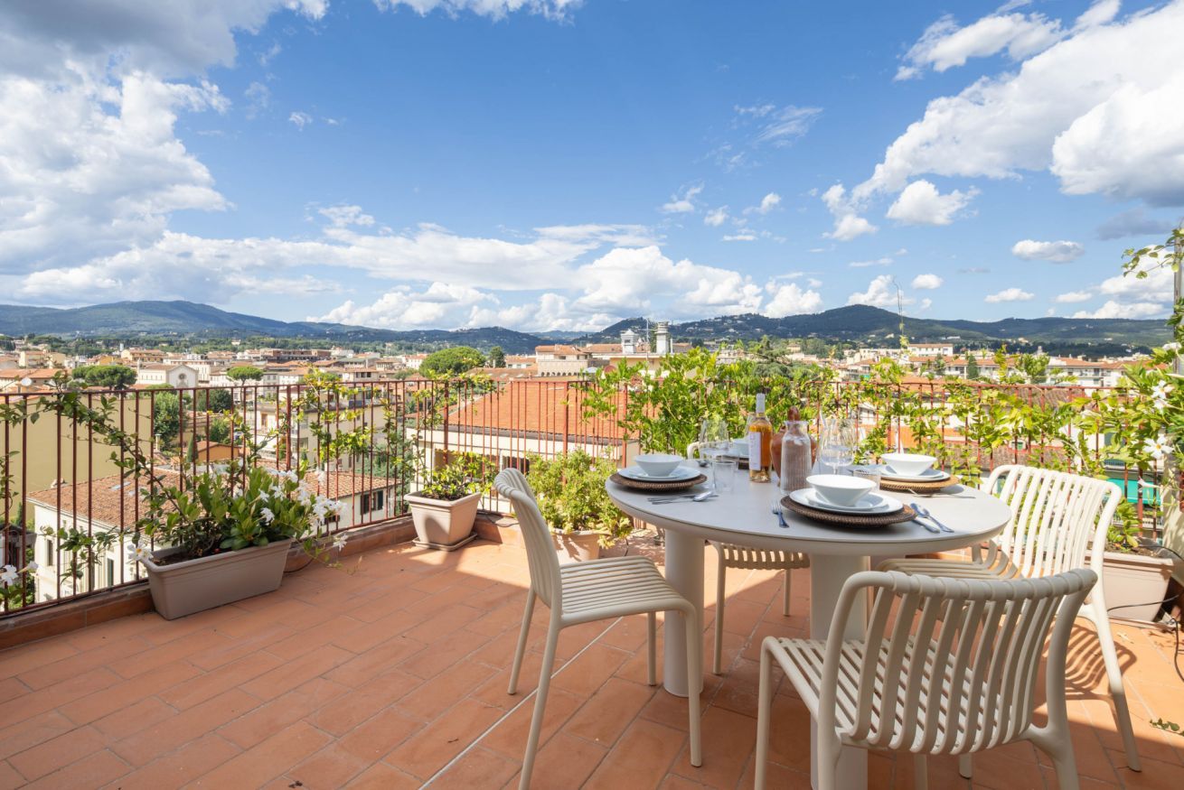 A terrace with a round table and chairs, potted plants, and a view of a cityscape with mountains in the background.