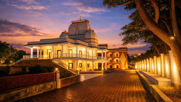 A large, multi-story building with a central dome and multiple balconies, surrounded by a paved walkway and trees. - Gangabagh by Divine Trails - Hotel Near Har Ki Pauri, Haridwar (Haridwar)