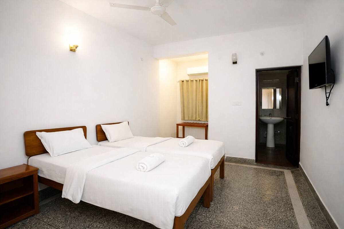 A hotel room with two beds, a bedside table, a TV, a bathroom with a sink, and a ceiling fan.