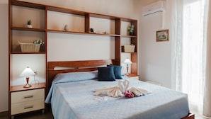 A hotel room with a bed, wooden headboard, bedside tables with lamps, a shelf with decorative items, and a window with curtains.