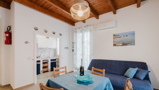 A room with a dining table, chairs, a sofa, a chandelier, a fire extinguisher, and a painting on the wall.