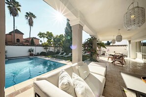 Ein Poolbereich mit weißer Couch, hängenden Laternen und Blick auf Pool und Palmen.