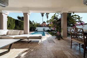 Un espacio al aire libre techado con piscina, un sofá blanco y una silla de madera.