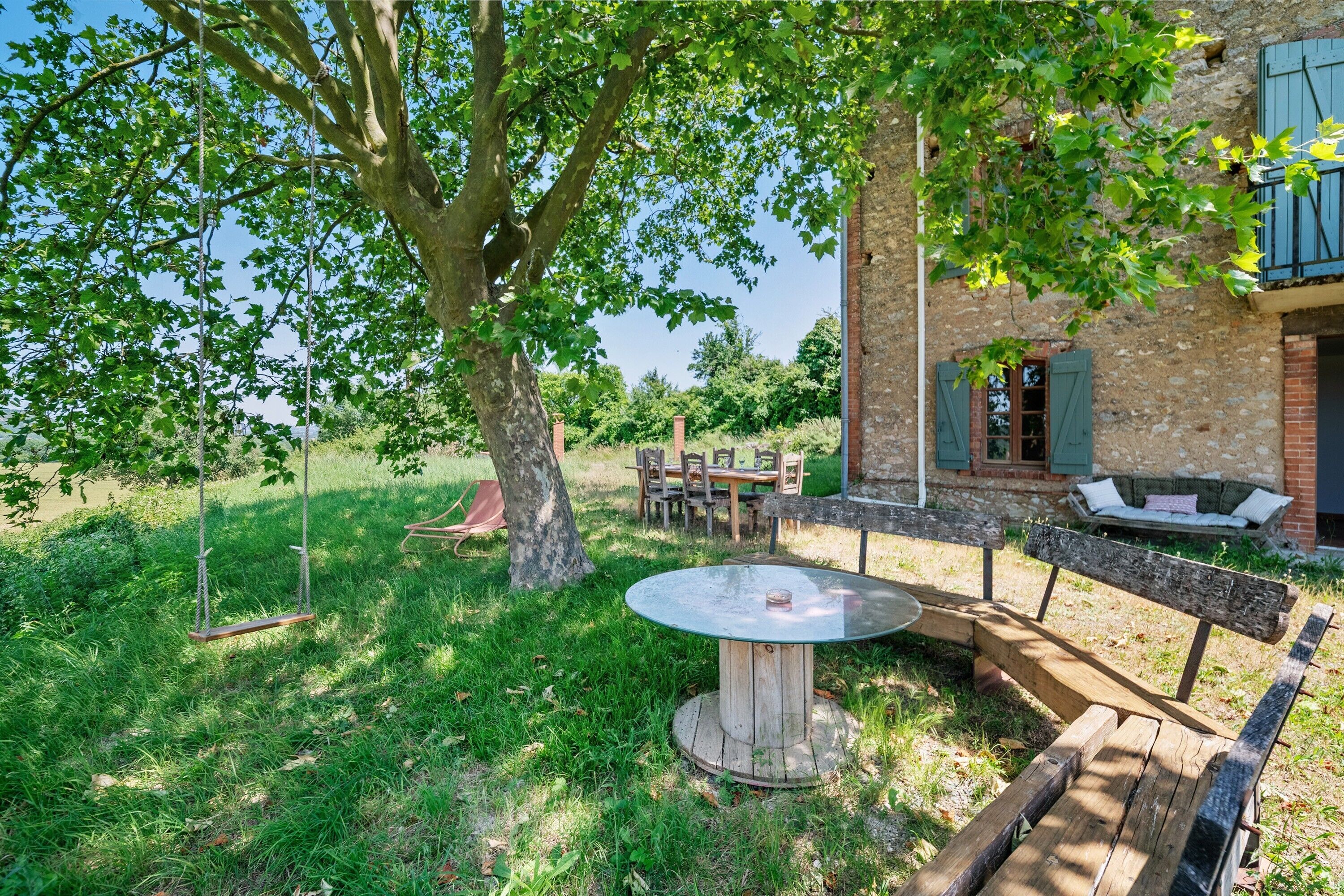 Een rustieke buitenzitplaats met een ronde tafel en bankjes, omringd door weelderige begroeiing en een grote boom.