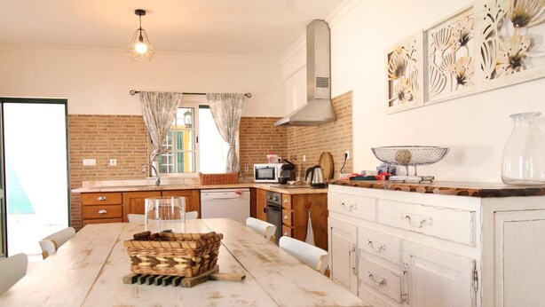 A kitchen with a brick wall, white cabinets, a dining table with chairs, and a hanging light fixture. - Villa GrandVale (Castro Marim)