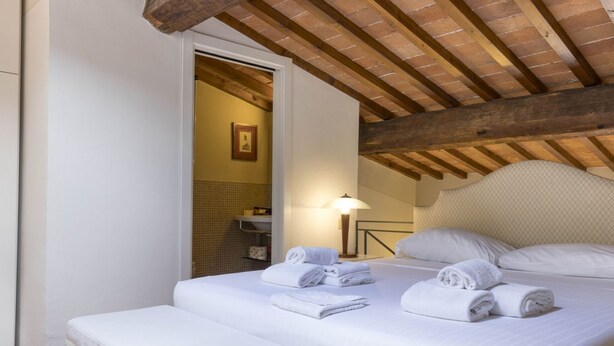 Una cama bien hecha con sábanas y toallas blancas, techo de madera y una pequeña mesita de noche con lámpara.