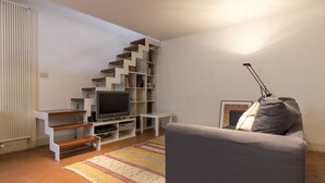 Una sala de estar con un sofá gris, un televisor de pantalla plana, una escalera con estanterías y una alfombra estampada.