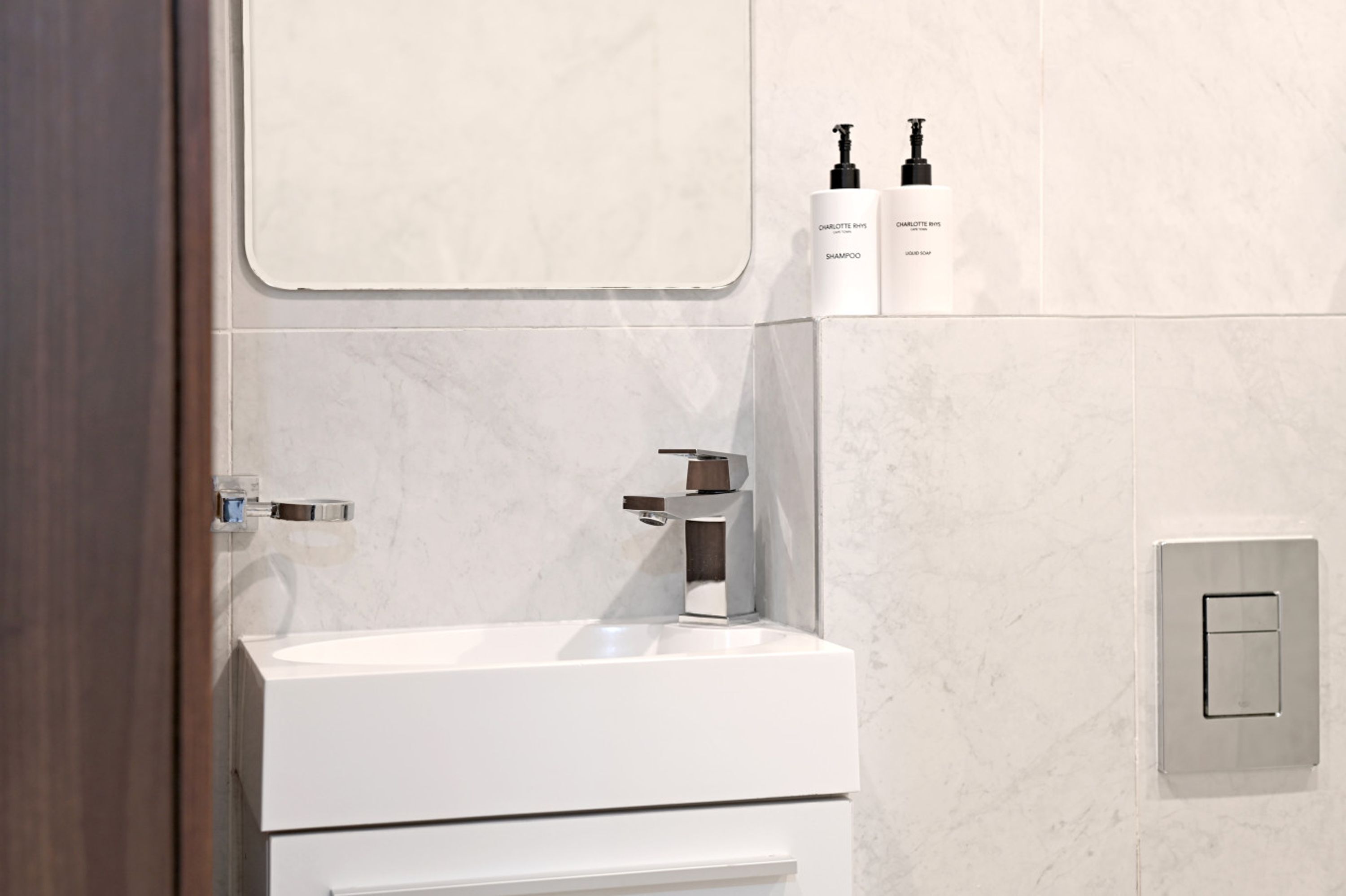 Ein modernes Badezimmer mit einem weißen Waschbecken, einem Spiegel und zwei Flaschen Charlotte Riviere Shampoo.