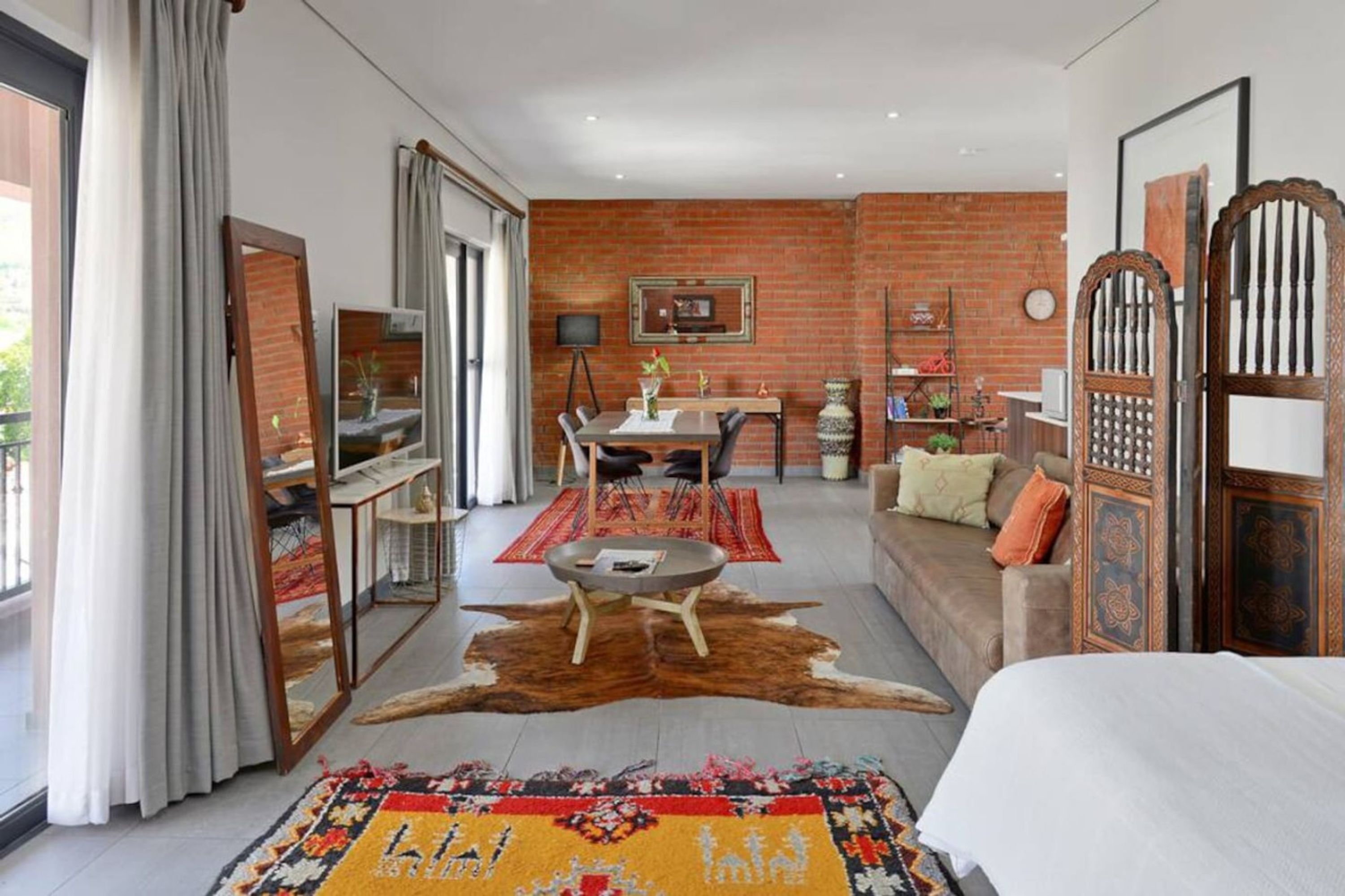 A modern hotel room with a brick wall, a dining area with a table and chairs, a sofa, a bed, and a large mirror.