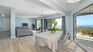 A modern living room with a dining table, grey sofa, and large windows overlooking a pool and ocean. - Spacious Family Camps Bay Escape Spectacular Views Pool Garden (Cape Town)