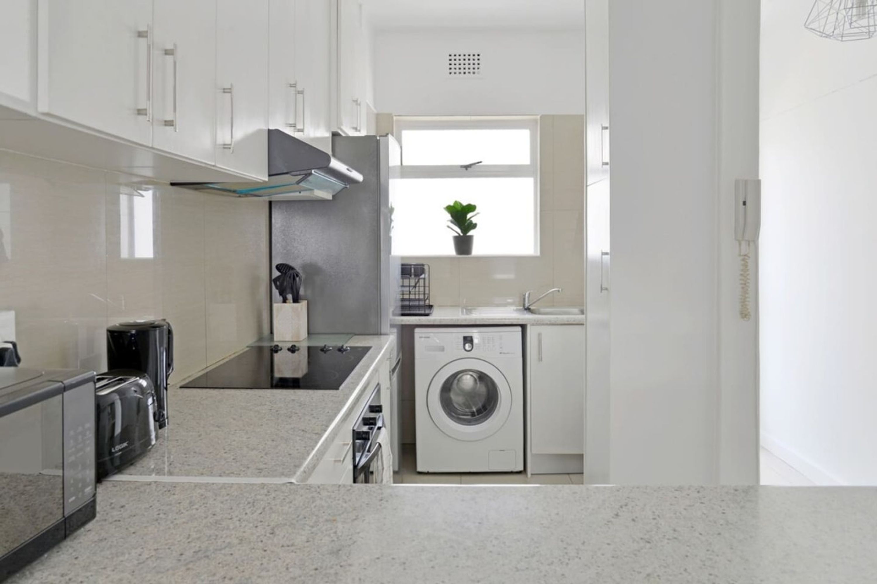 A compact kitchen with a washing machine, a coffee maker, and a window with a plant.