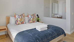 A modern bedroom with a bed, colorful pillows, a bedside table with a lamp, and a bathroom with a mirror and sink. - Modern 1-bed V A Waterfront Canal Harbor Views (Cape Town)