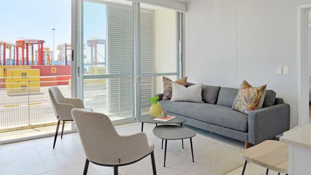 A modern living room with a grey sofa, two armchairs, a coffee table, and a view of a port with shipping containers. - Modern 1-bed V A Waterfront Canal Harbor Views (Cape Town)