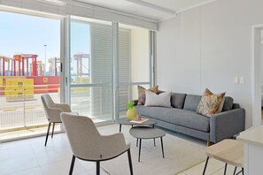 Una sala de estar moderna con un sofá gris, dos sillones, una mesa de centro y vista a un puerto con contenedores de carga.