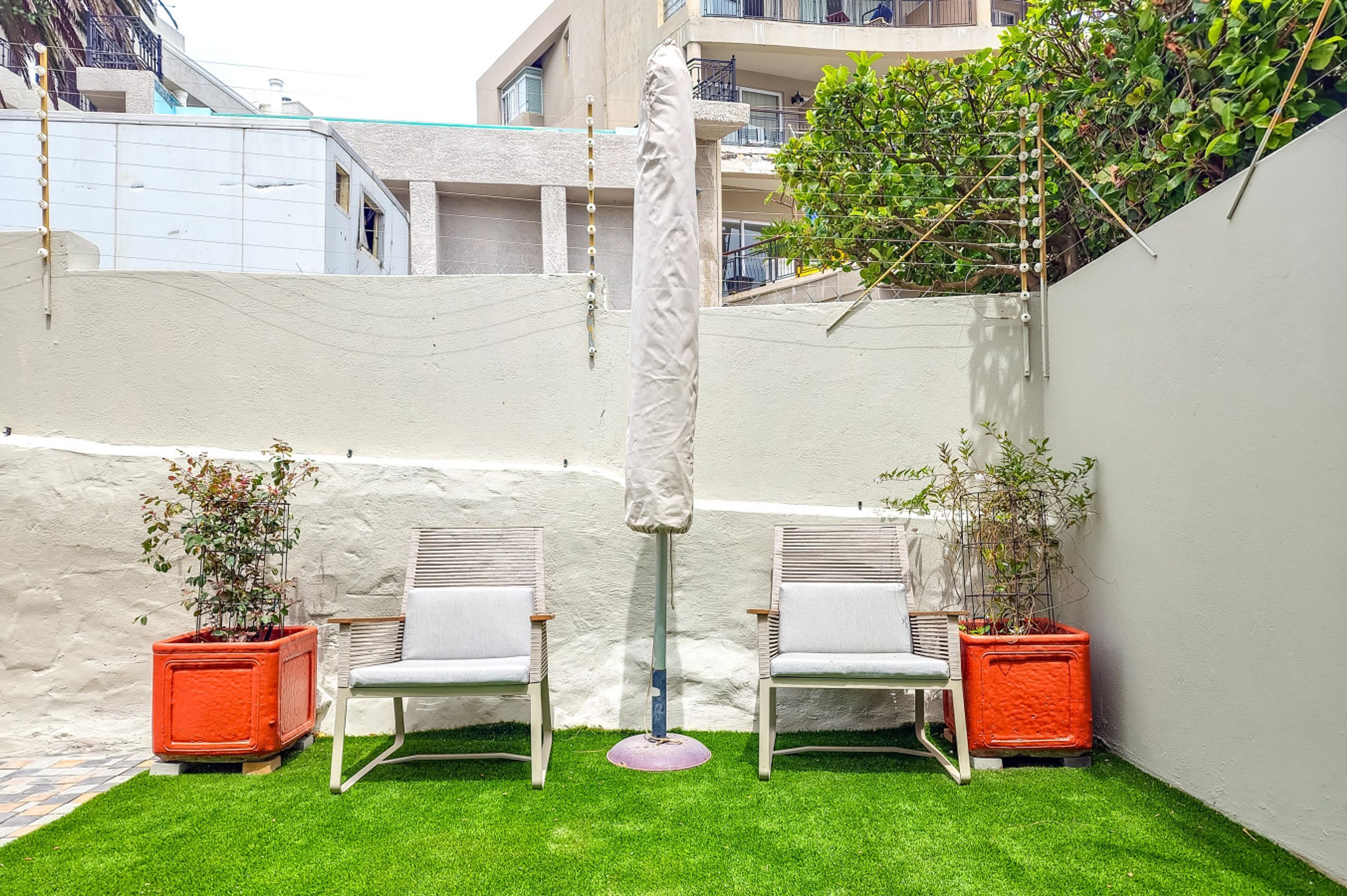 A small outdoor seating area with two white chairs and a table, surrounded by potted plants and a white wall.